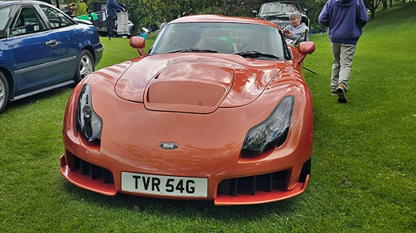 Front view of an orange TVR Sagaris sports car displaying its prominent "TVR 54G" UK private number plate on grass.