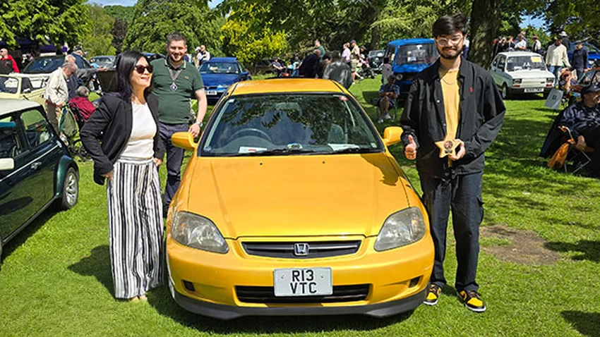 Yellow Honda Civic with private plate R13 VTC at a car show, owner proudly holding a trophy.