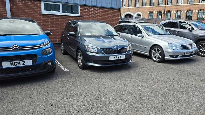 Several cars parked, featuring UK private number plates: MGM 2, XFVI, and NEN 555 on a blue Citroen, grey Skoda, and silver M